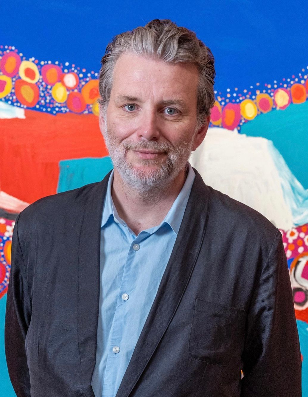 Man in blue shirt and black jacket smiles in front of a colorful abstract painting.