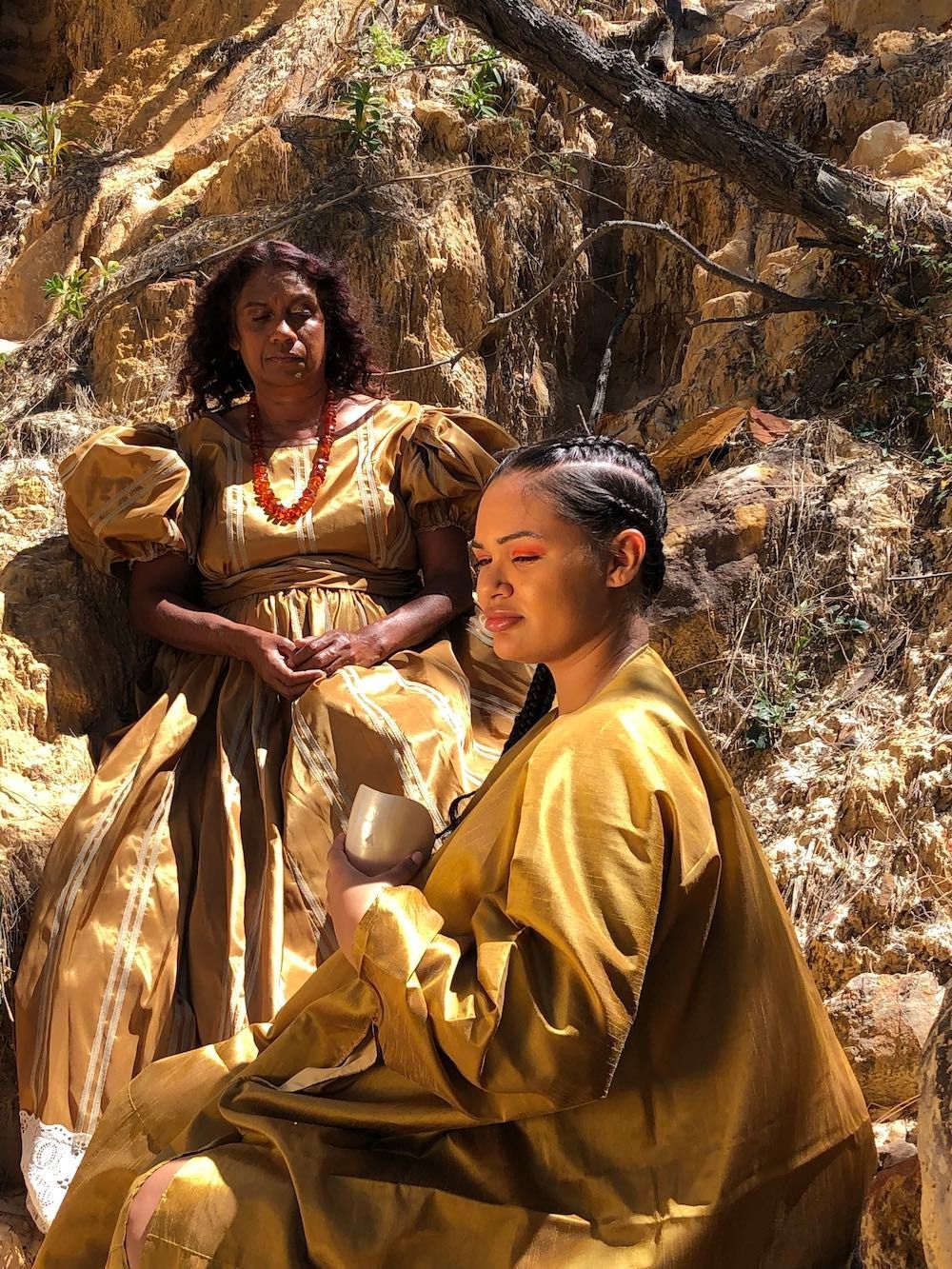 Two people in golden, textured garments sit among rocky, sunlit terrain, with one holding a small, metallic vessel.