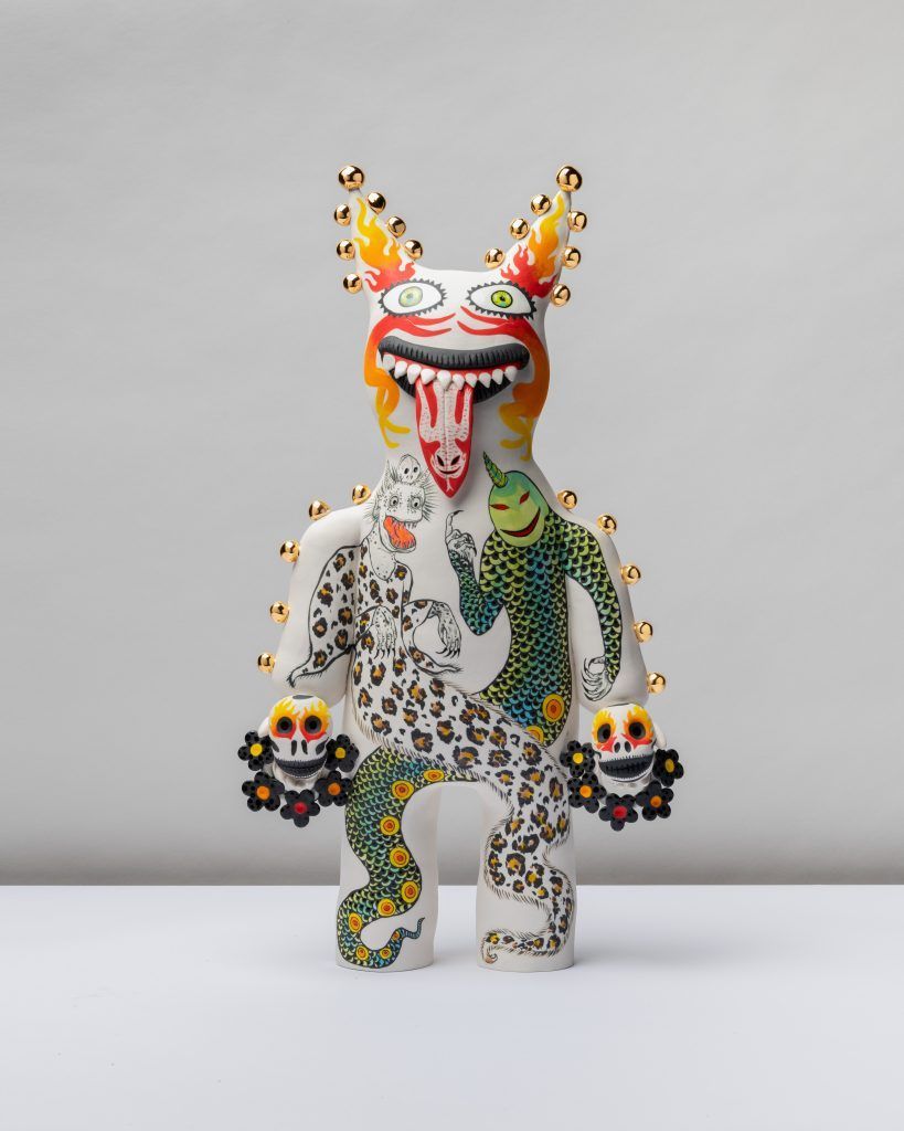 A stylized, white humanoid sculpture with a monstrous, red-tongued face, patterned body, and two small skulls for hands.
