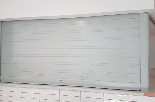 A closed, light gray horizontal slat rolling security shutter installed above a tiled counter.