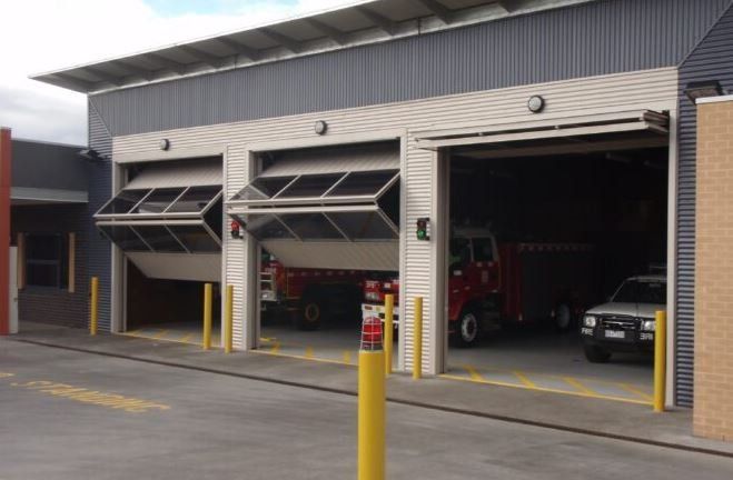 A fire station exterior with three large bays, two featuring bifold garage doors opening upward, revealing fire trucks.