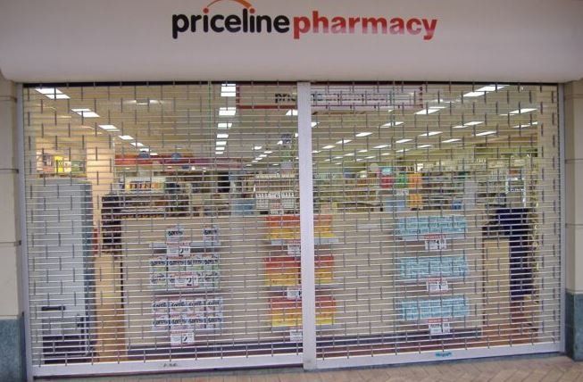A closed Priceline Pharmacy storefront viewed through a metal security gate.