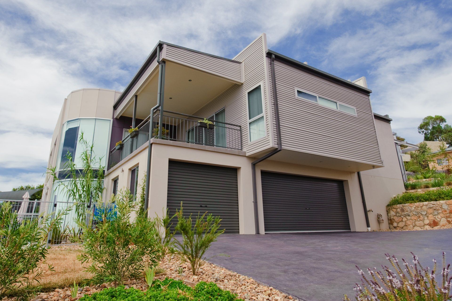 A two-story modern house with a beige exterior, a metal-sided upper level, a balcony, and two dark garage doors.