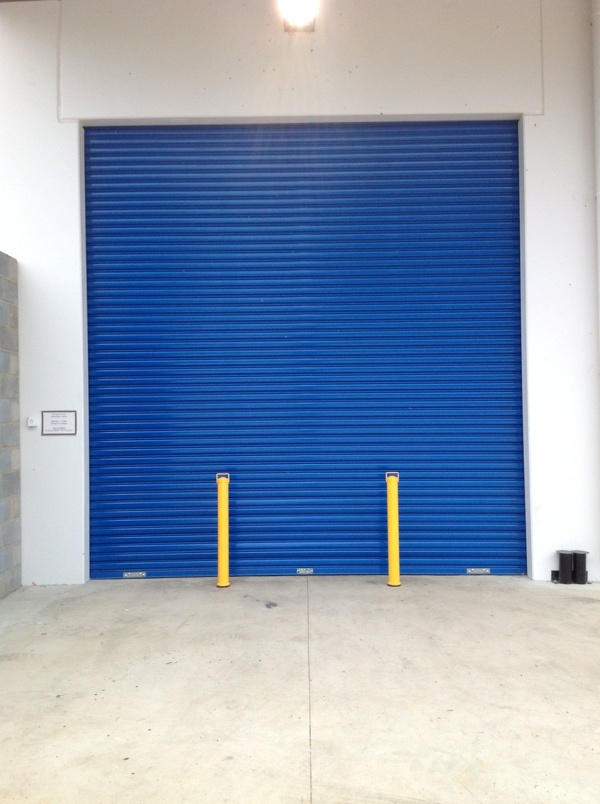 Blue roll-up metal door with two yellow safety bollards in front of it on a concrete surface.