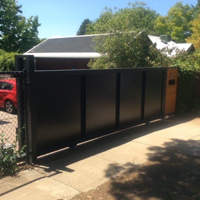 A modern black sliding metal gate with vertical supports and a wood-paneled side section near a paved driveway.