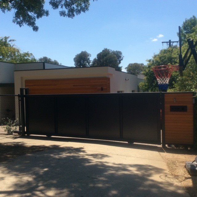 A black sliding gate with wood accents in front of a modern home, next to a basketball hoop on a sunny day.