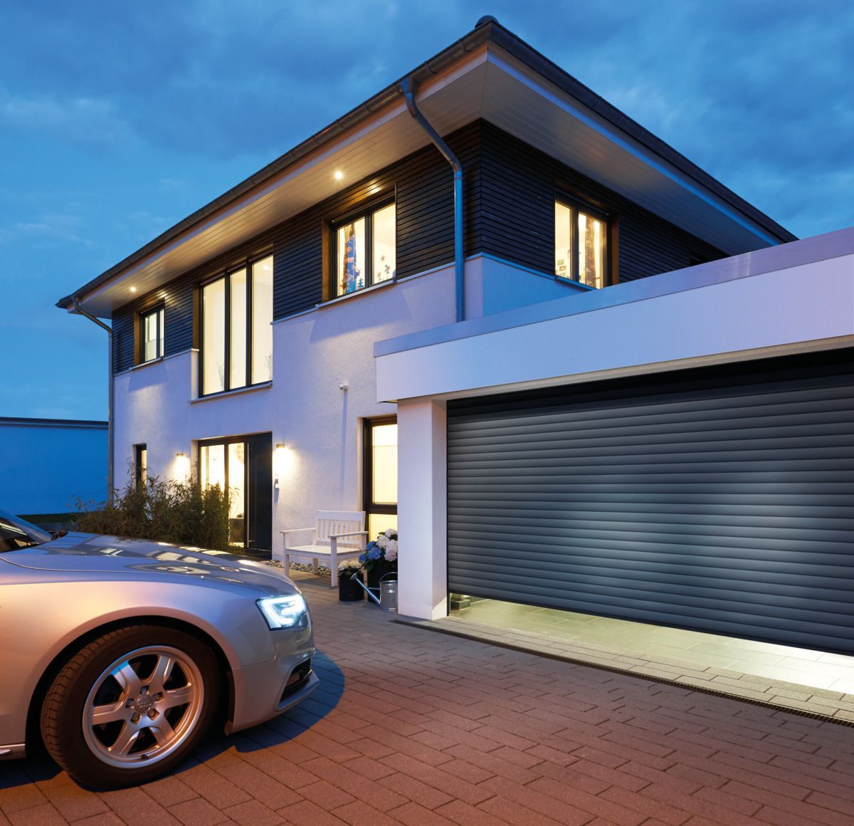 A silver car parked in front of a modern two-story home with a dark gray rolling garage door at dusk.
