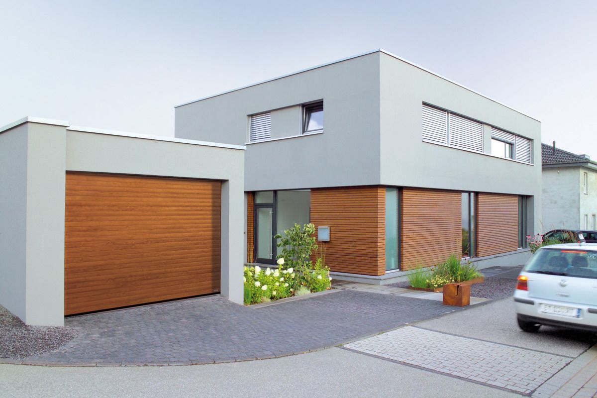 A modern two-story home with light gray walls, wooden cladding, a garage, and a small garden, seen from a paved driveway.