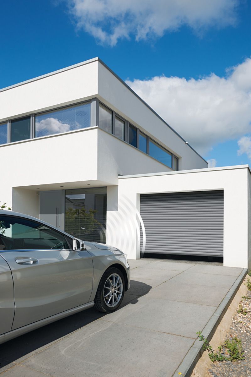 A modern white house with a driveway and a gray car parked in front of a closed metallic garage door under a blue sky.