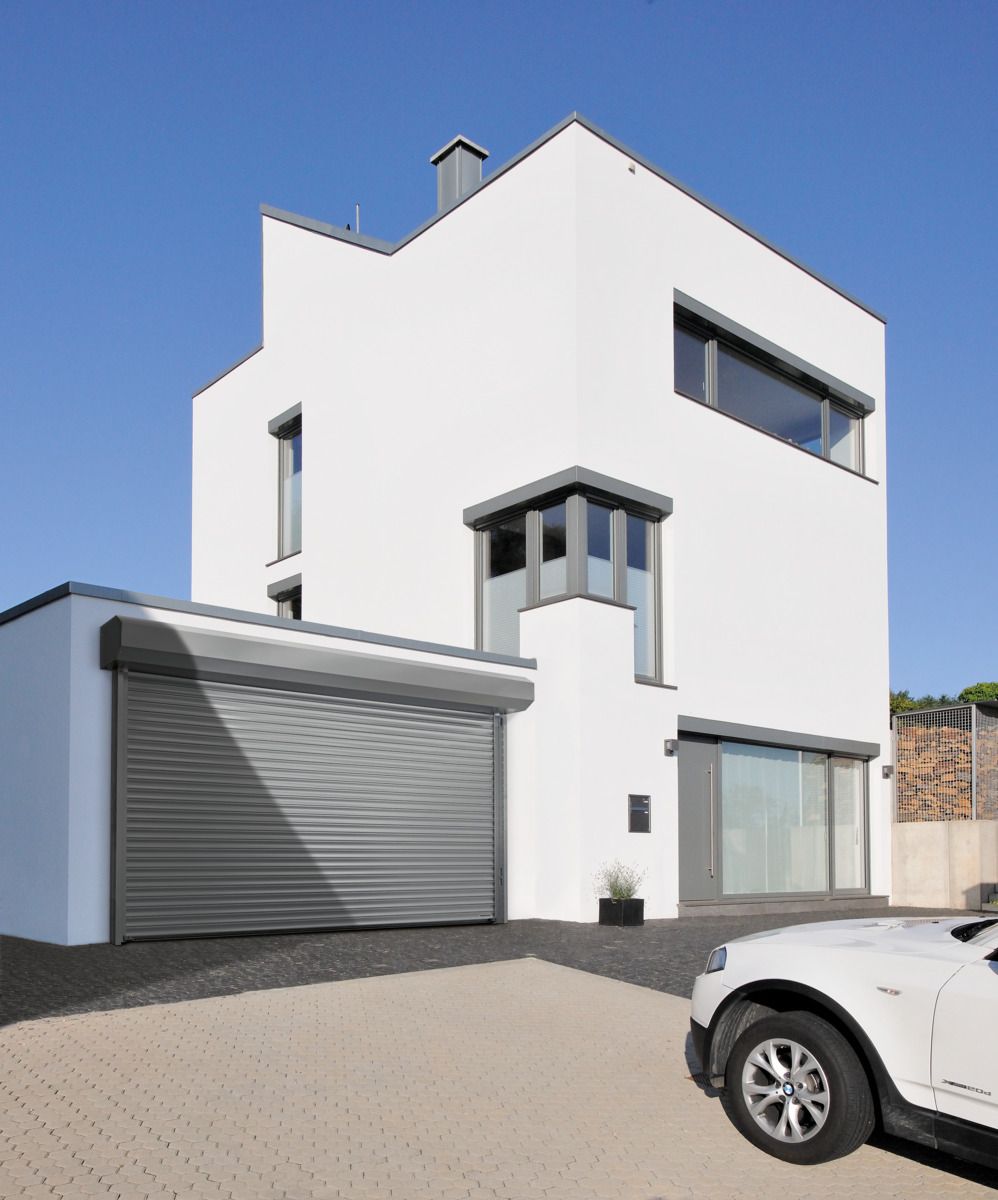 A modern white house with a gray rolling garage door, corner windows, and a car parked on a paved driveway under blue sky.