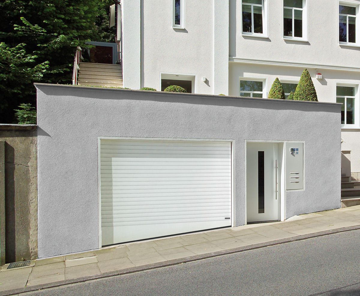 A gray building facade featuring a white, horizontally paneled garage door next to a white door on a sloped street.