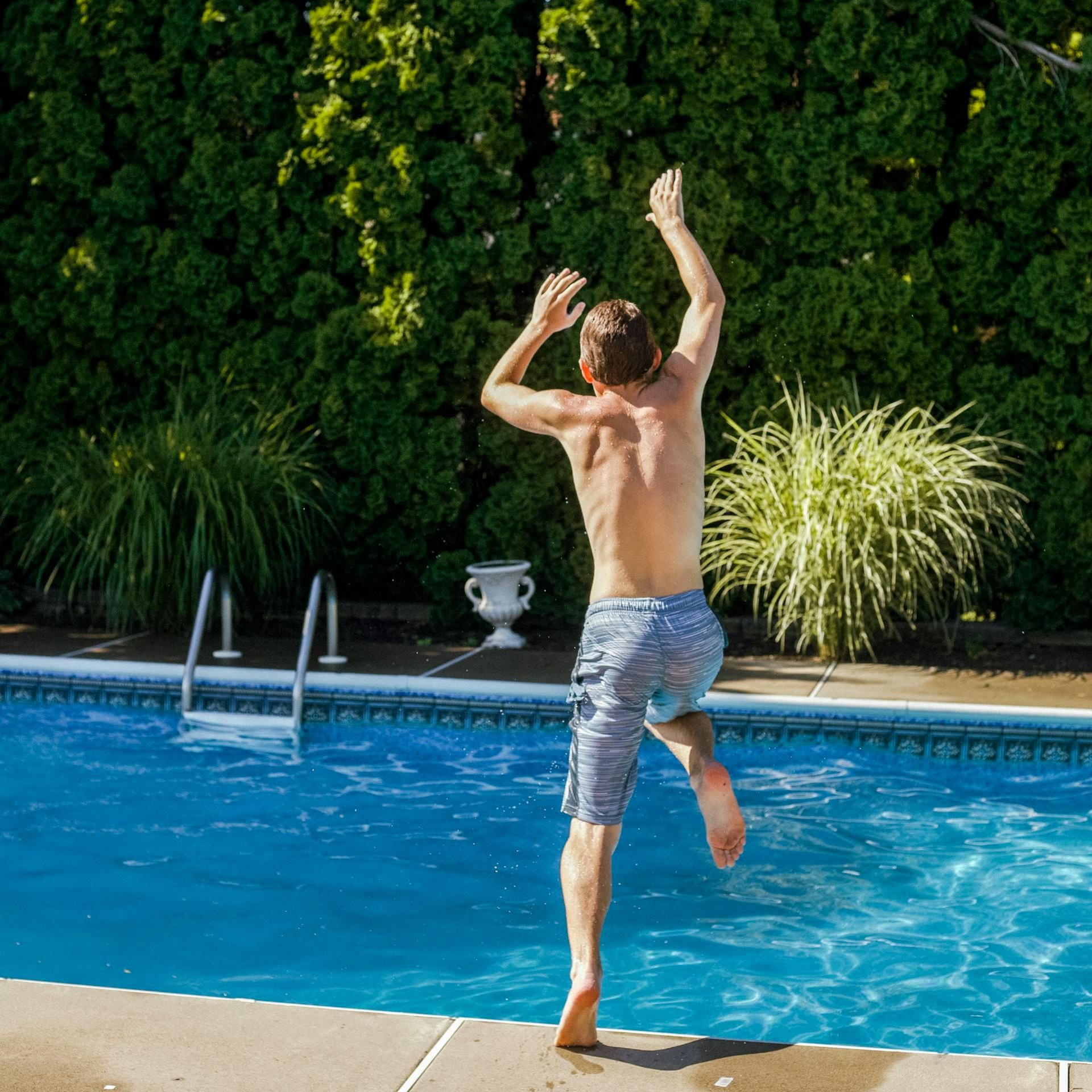 Man jumps into a pool with arms raised, blue water and shorts, green bushes in the background at Fusion Apartments in Irvine CA  at Fusion Apartments in Irvine CA.