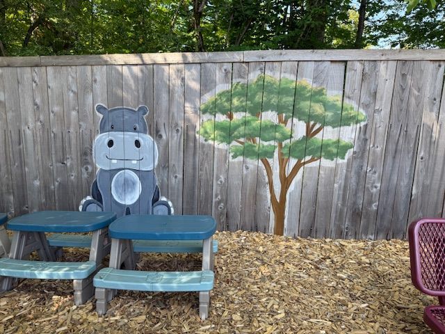 Wooden fence with painted hippo and tree; two small blue tables in front.