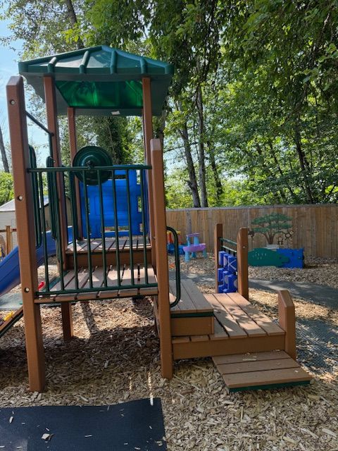 Playground with wooden structure, blue slide, and climbing features, set on wood chips, backed by trees.