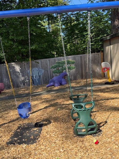 Playground with three swings, wooden chips, a fence, and a purple hippo statue.