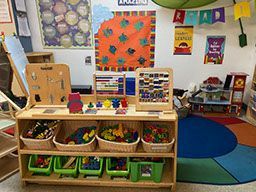 Wooden shelf with toys in a classroom setting, a colorful rug and learning posters visible.