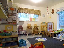 Daycare room, small chairs, high chairs, cribs, window, toys, and bulletin board.