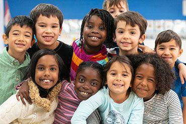 Group of smiling children, diverse ethnicities, posing close together in a well-lit indoor setting.