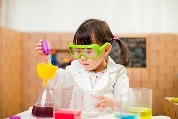 Young child in lab coat and goggles pouring liquid into a beaker, surrounded by colorful beakers.