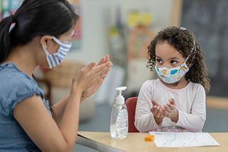 Teacher and child wearing masks use hand sanitizer in a classroom.