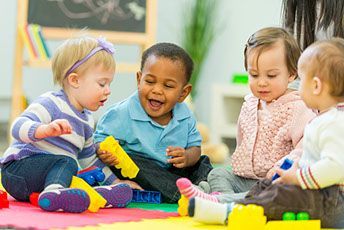 Four toddlers playing with toys on a colorful mat; one child laughs.