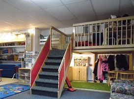 Playroom with a loft. Stairs with red sides lead to a wooden platform. Below is a coat rack and small table.