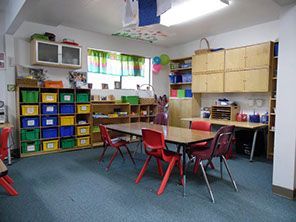 Preschool classroom interior with tables, chairs, storage bins, and cabinets.