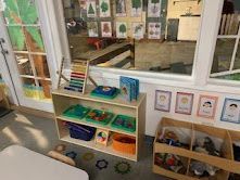 Shelves with educational toys and materials in a brightly lit classroom, window with art on display.