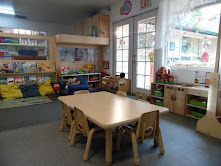 A brightly lit classroom with a table and chairs, shelves, toys, and a loft bed near a window.