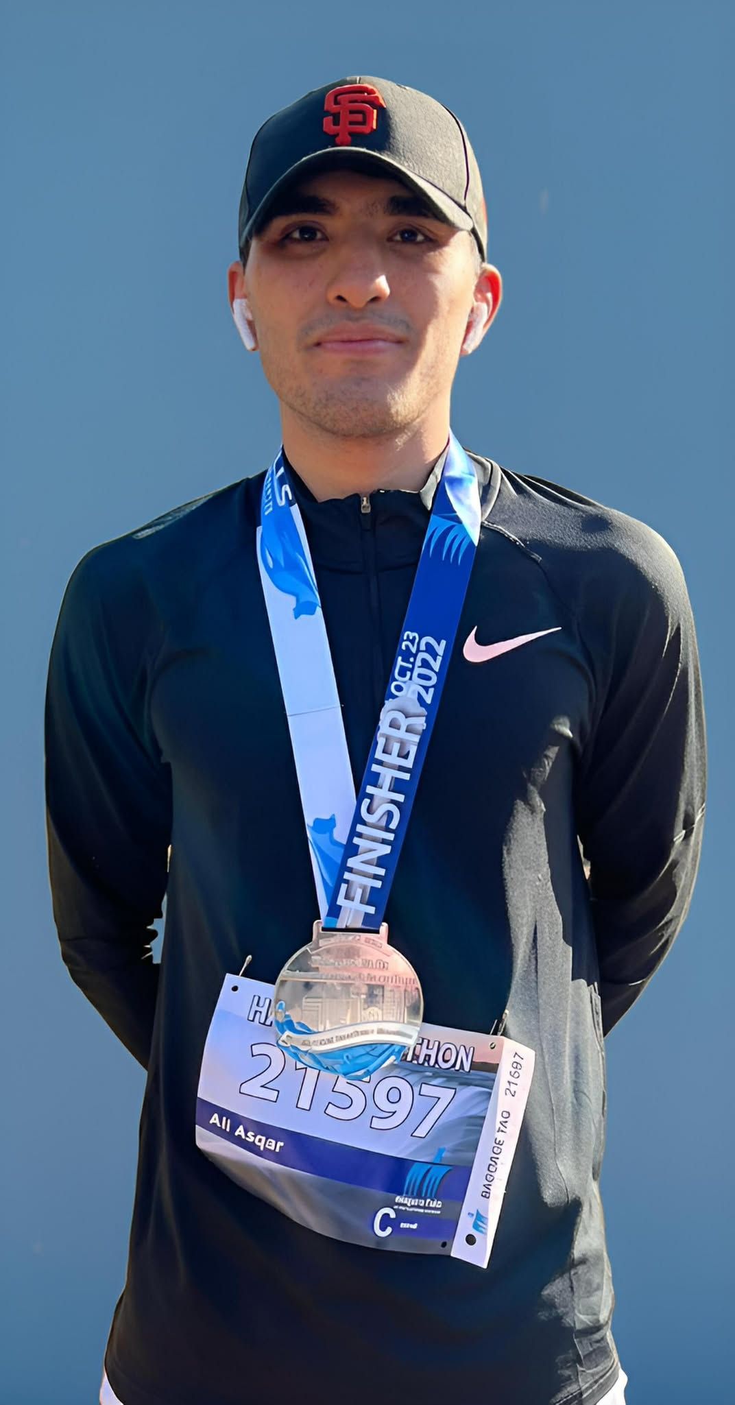 Man wearing a San Francisco Giants hat, medal, Nike shirt, posing against a blue background.