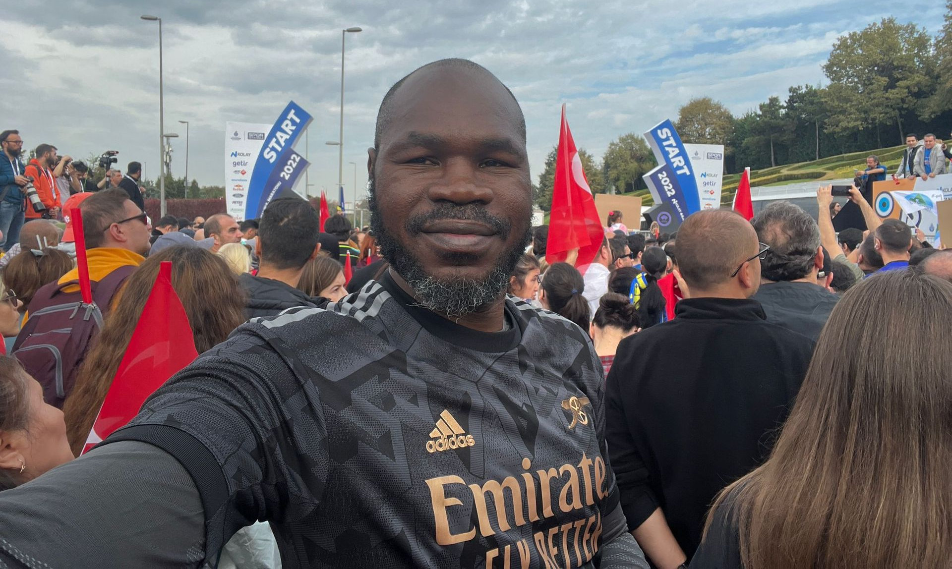 Man in jersey smiles at camera, surrounded by crowd holding signs with red flags.