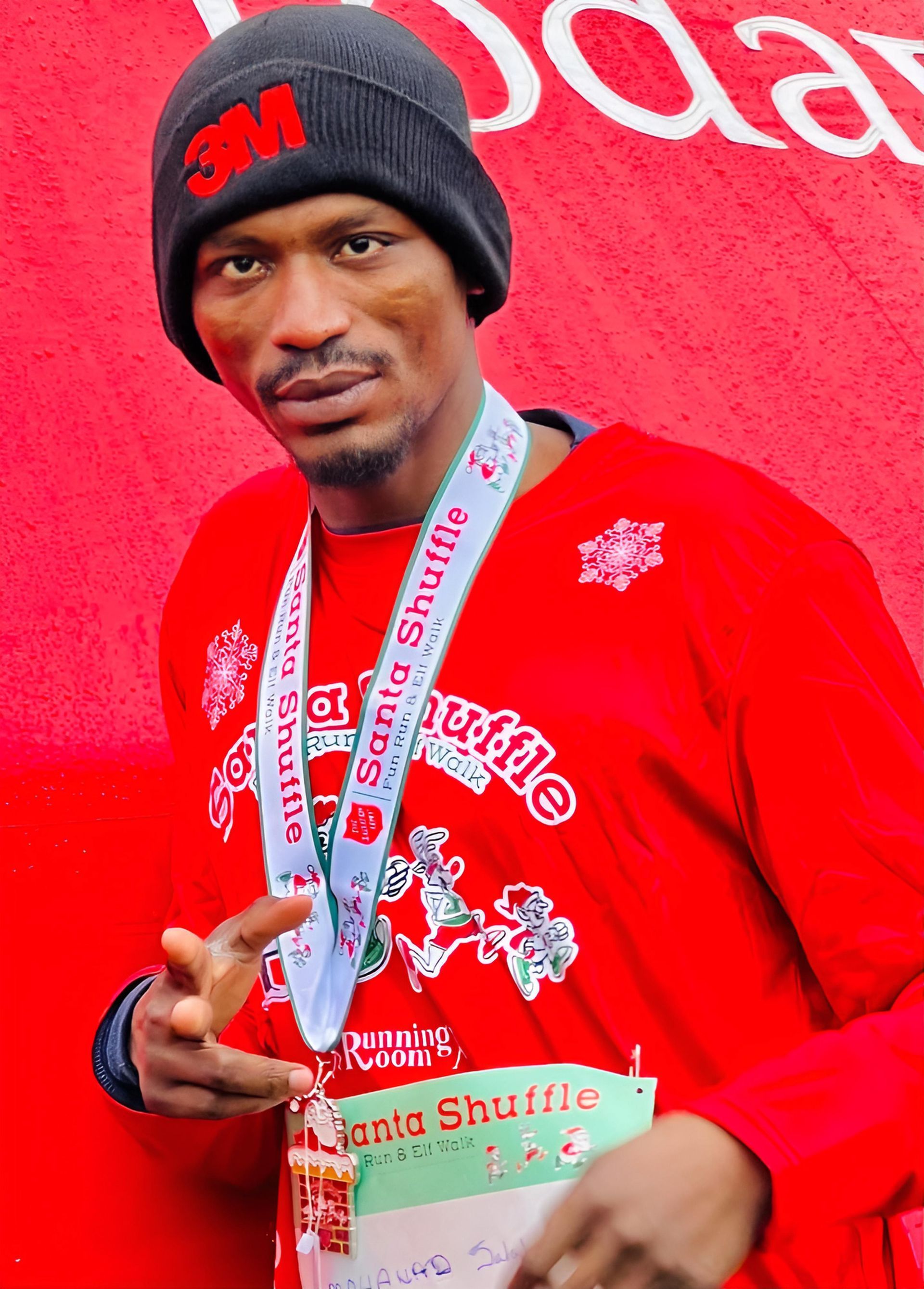 Man wearing a red Santa Shuttle shirt, beanie, and medal, smiling and making a peace sign.