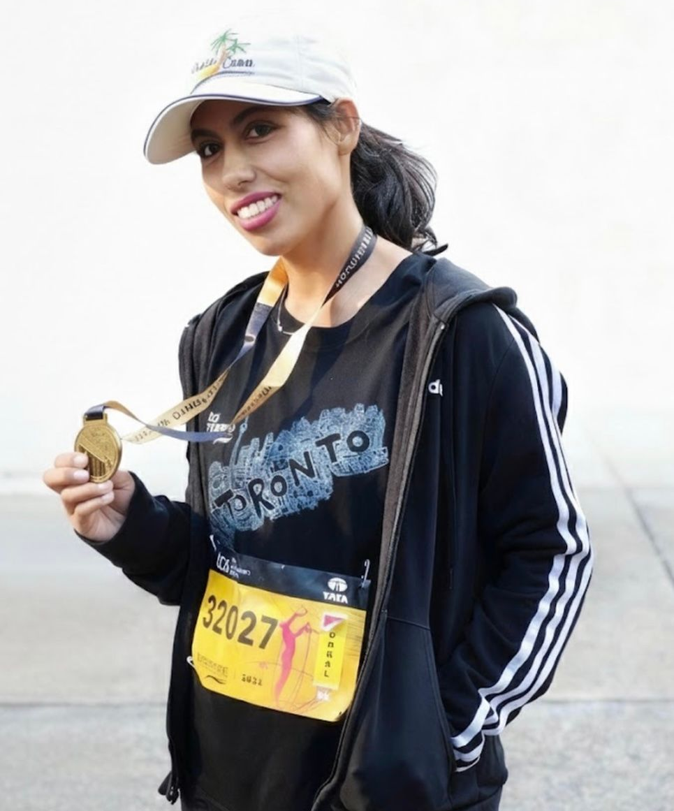 Woman wearing hat, holding a medal, with a race bib, standing outside.