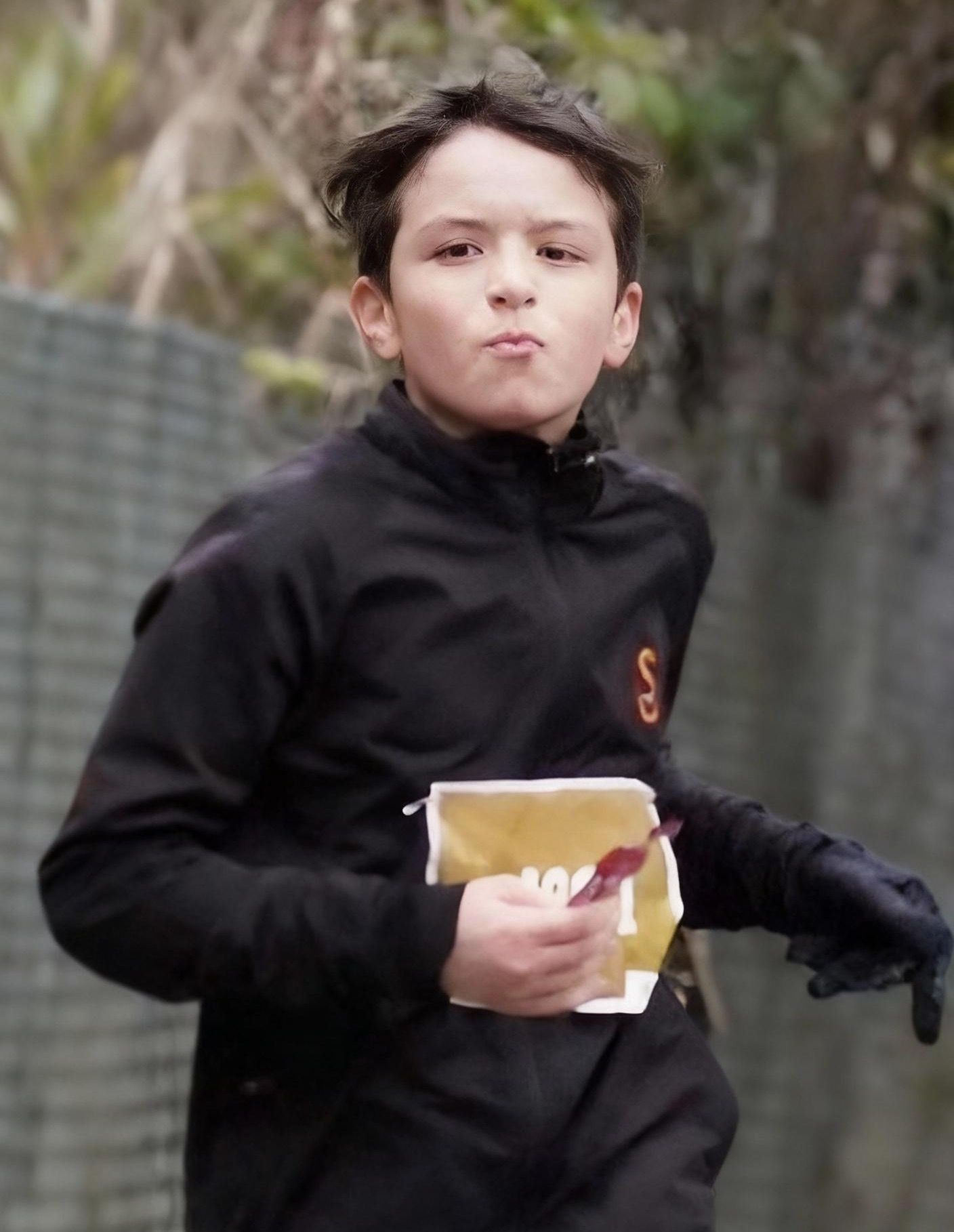 Boy running, holding a snack, in a black jacket with a badge, looking at the camera.
