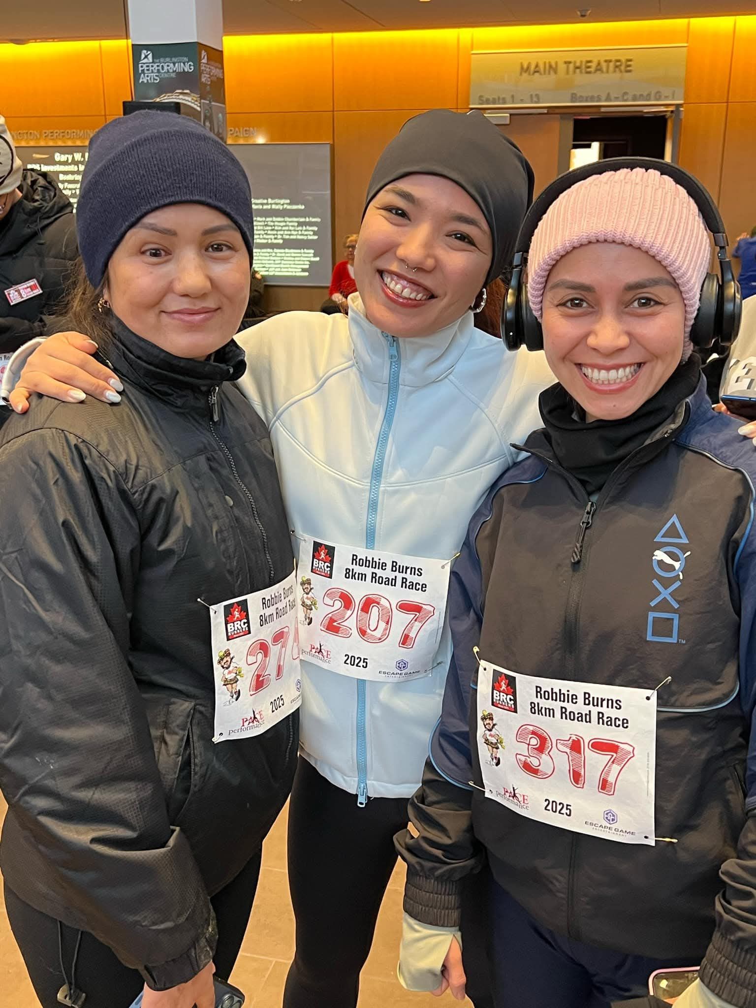 Three women in athletic wear, smiling and posing. Wearing bibs, beanies, and a headband. Indoors.