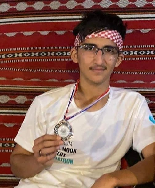 Teenager with glasses wearing a headband and holding a silver medal, smiling. Red patterned backdrop.