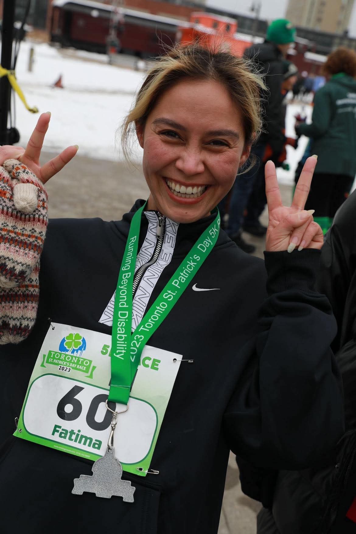 Woman with bib #60, a St. Patrick's Day race medal, makes peace signs, smiles, and wears a black jacket.