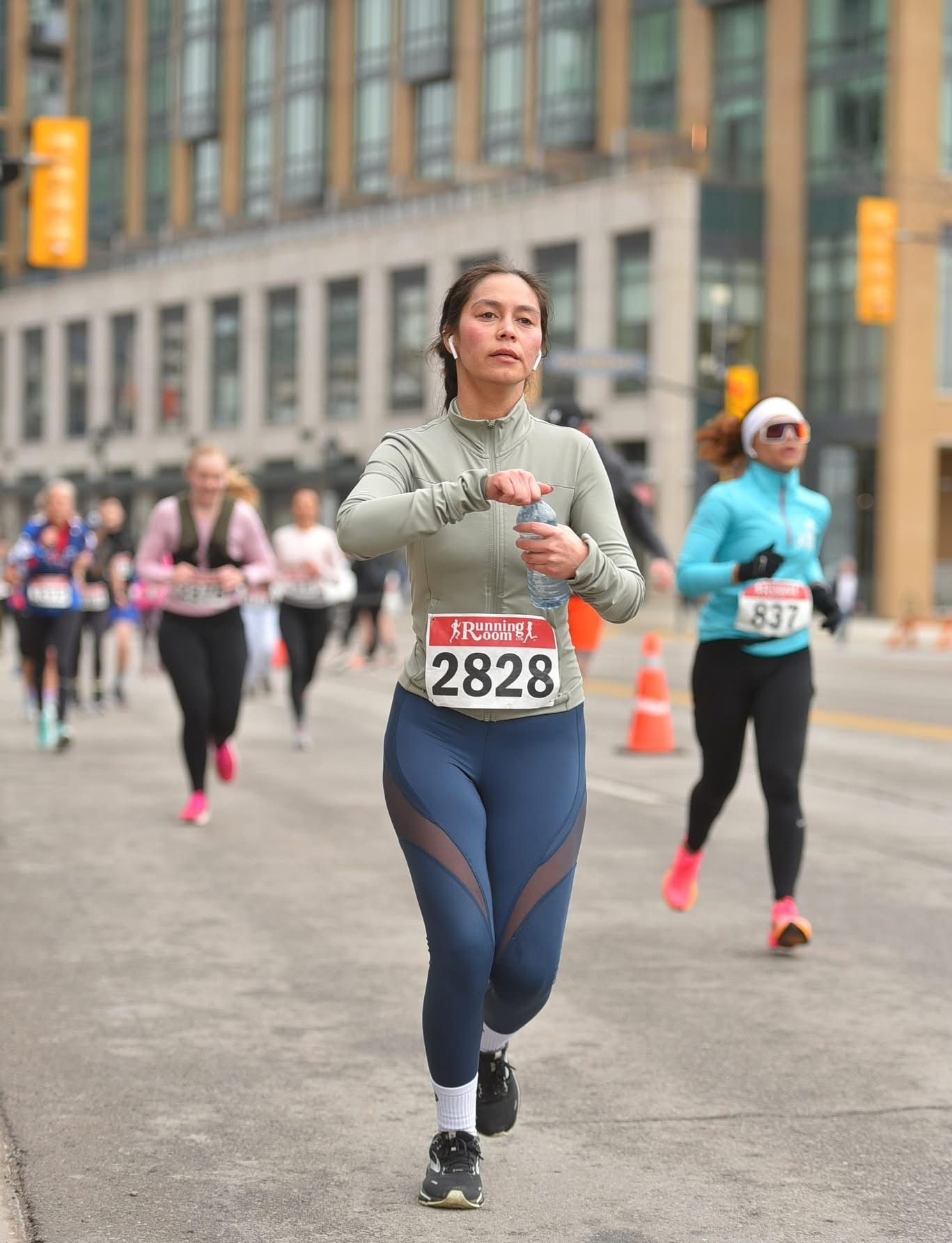 Woman running in a marathon, wearing a bib, holding water bottle, others run in background, urban setting.