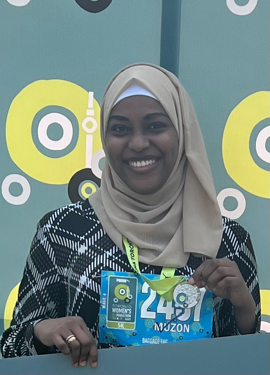 Woman in beige hijab smiles, holding medal and race bib with number 2431.