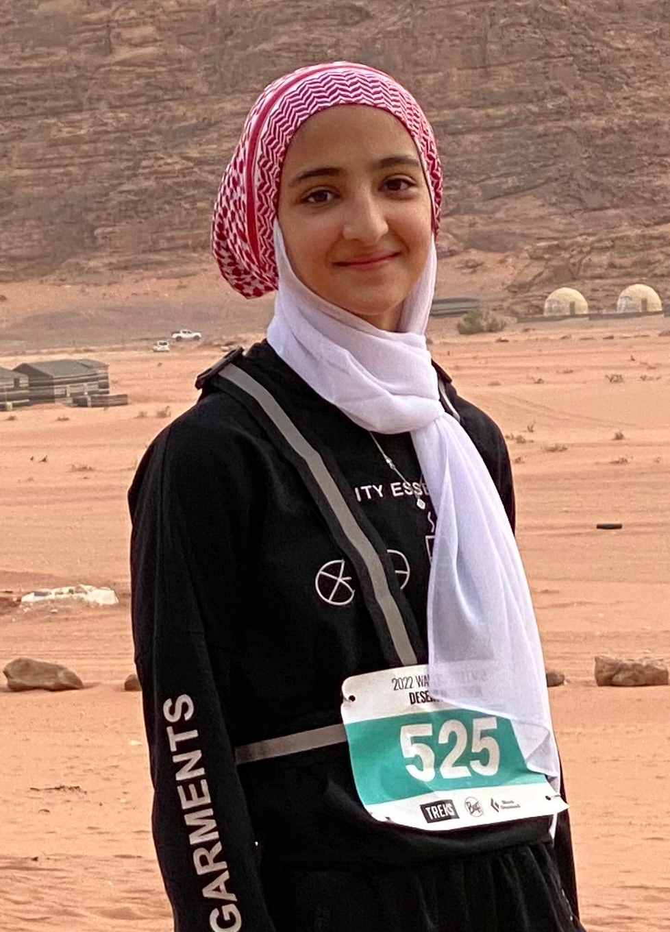 Woman in headscarf with race bib, smiling outdoors in desert landscape.