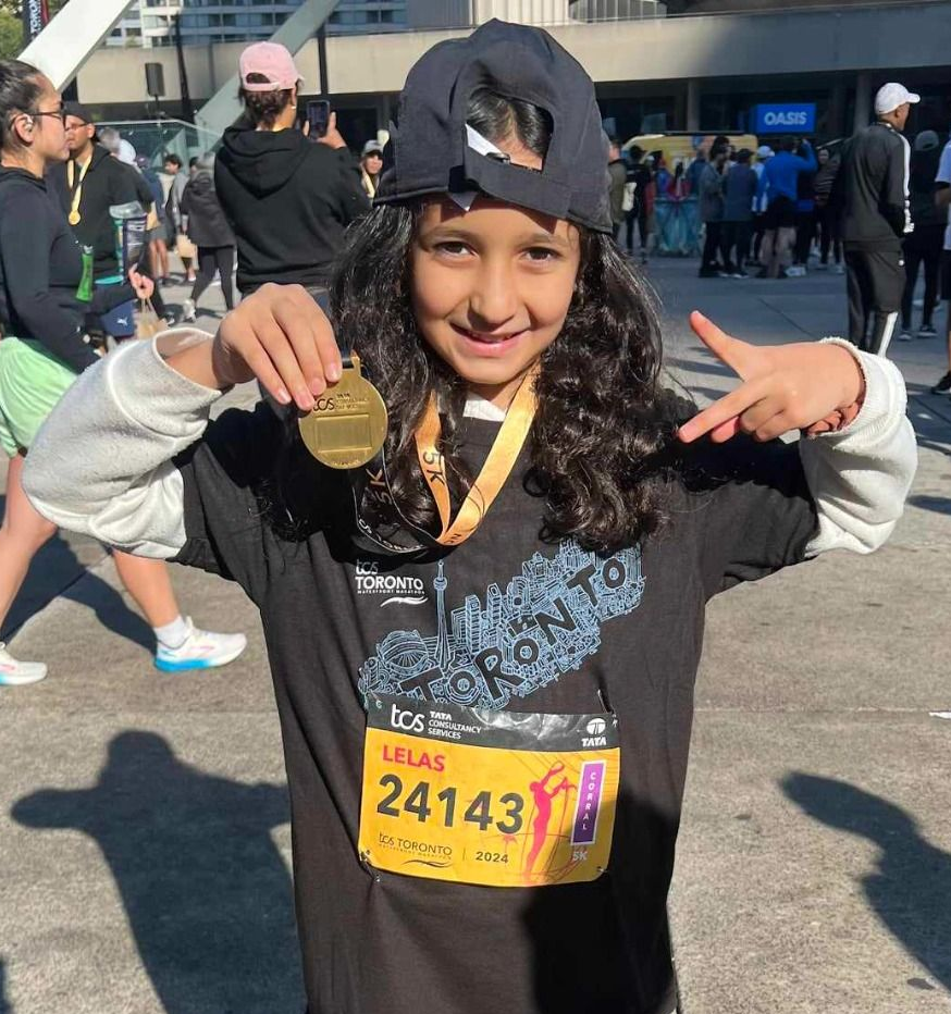 Young person with a medal, pointing, wearing a race bib and hat in Toronto.