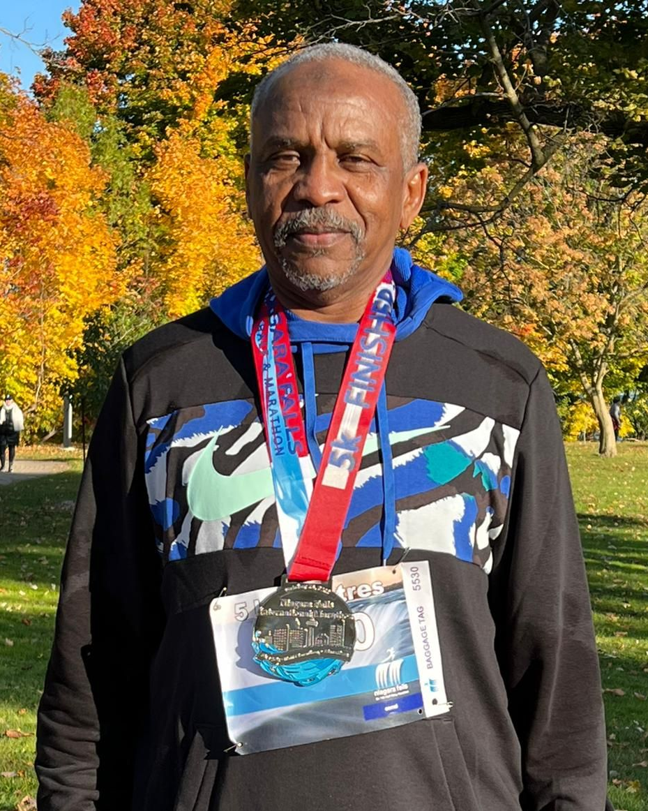 Man in black hoodie with medal outdoors. Fall foliage in background.