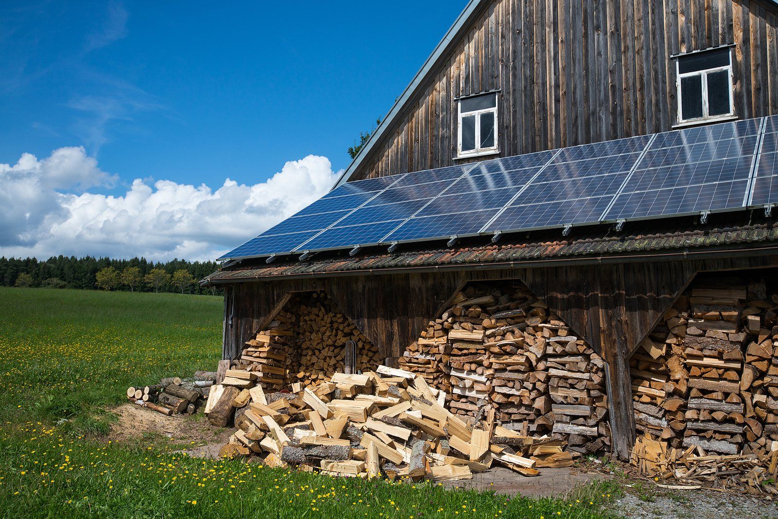 a wooden barn with solar panels on the roof and a pile of wood in front of it