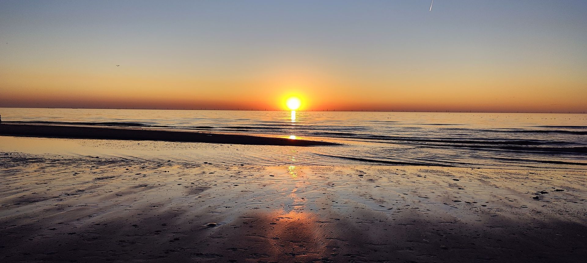 Zonsopgang boven een rustig strand, met oranje licht dat weerkaatst op het natte zand en de kalme golven.