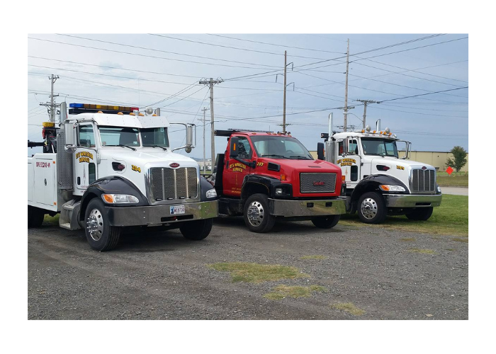 Three tow trucks parked in a gravel lot under cloudy sky, including white and red trucks.