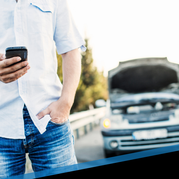 Man in white shirt holding phone beside broken-down car with open hood on roadside