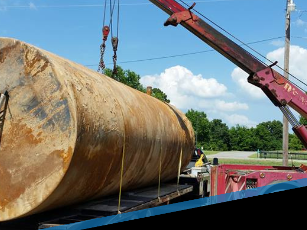 Large rusted cylindrical tank being lifted by a crane onto a flatbed truck outdoors