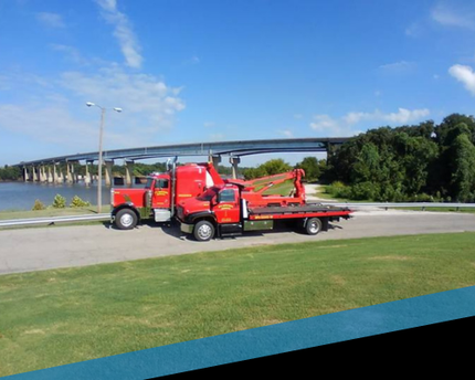 Red tow truck carrying a red tractor on a flatbed near a bridge by the water under a blue sky