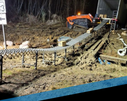 Orange excavator at muddy construction site beside a house, building a raised walkway with rebar forms.