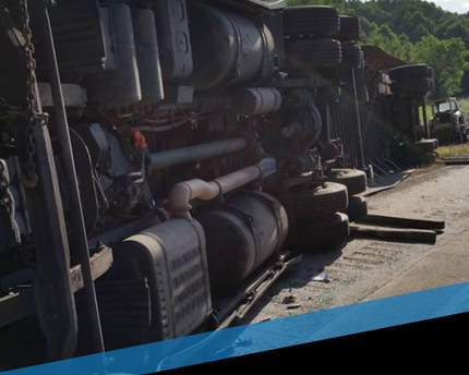 Overturned truck loaded with large cylindrical tanks on a dirt road beside greenery